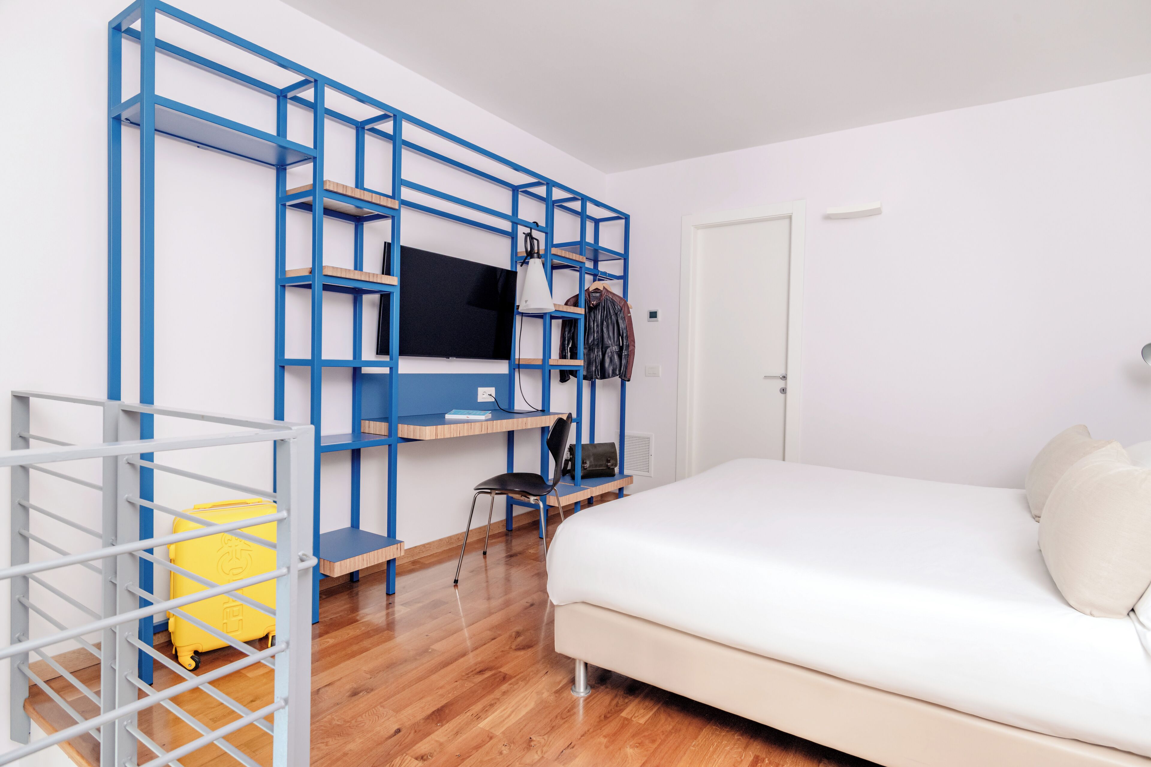 Una stanza con un letto, una TV montata su una mensola di metallo blu, una sedia e una valigia.