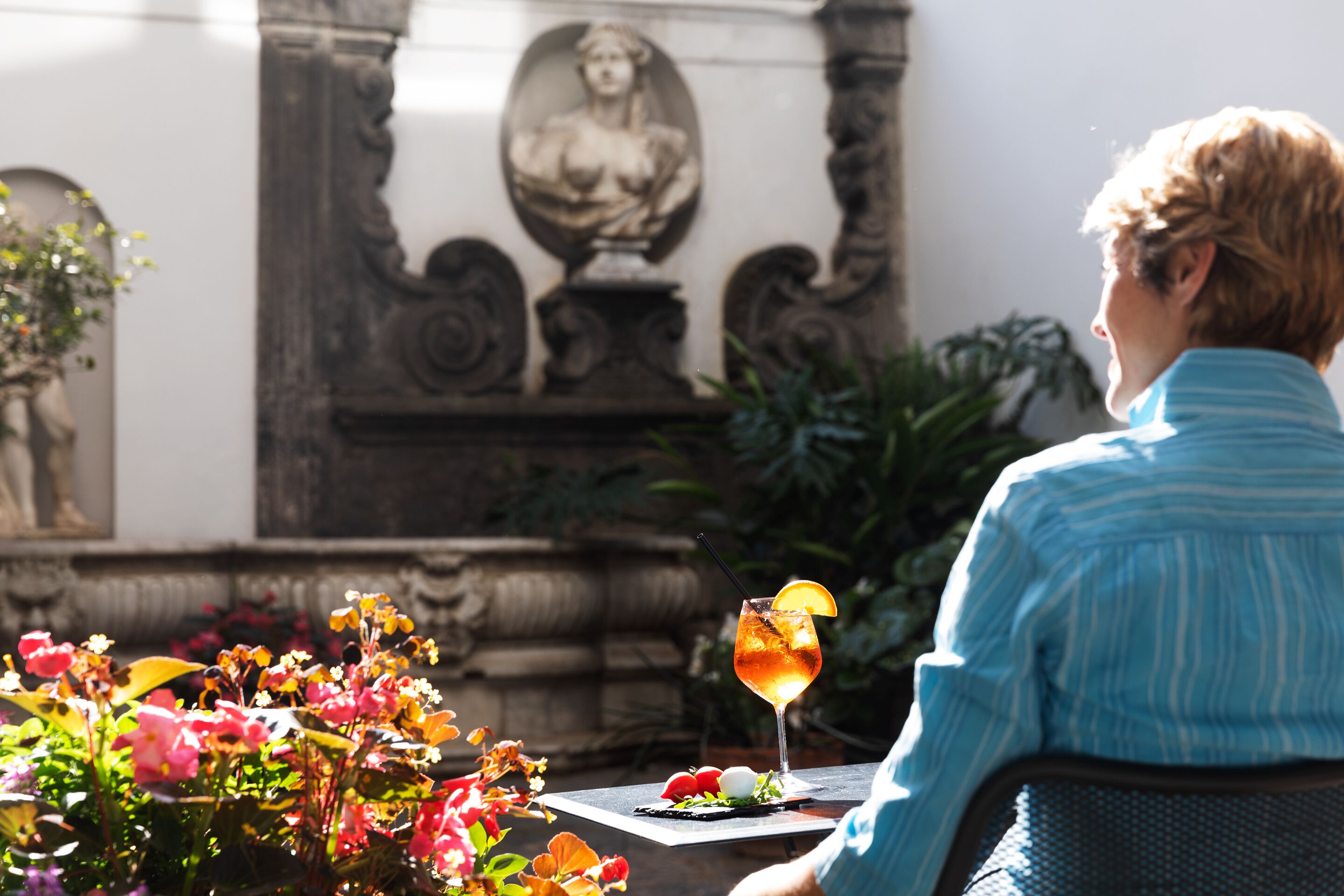 A person seated at a table with a drink, surrounded by flowers and a statue.