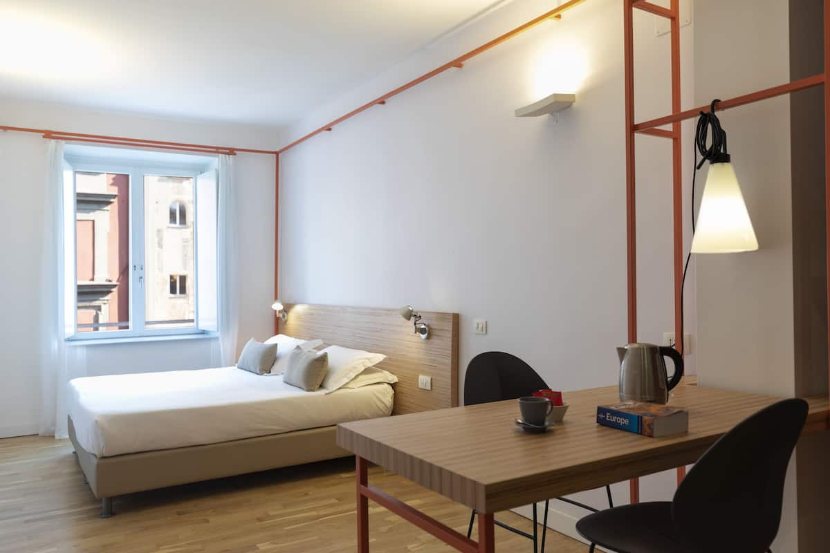 A bedroom with a bed, a table with a kettle, a chair, and a window with curtains.