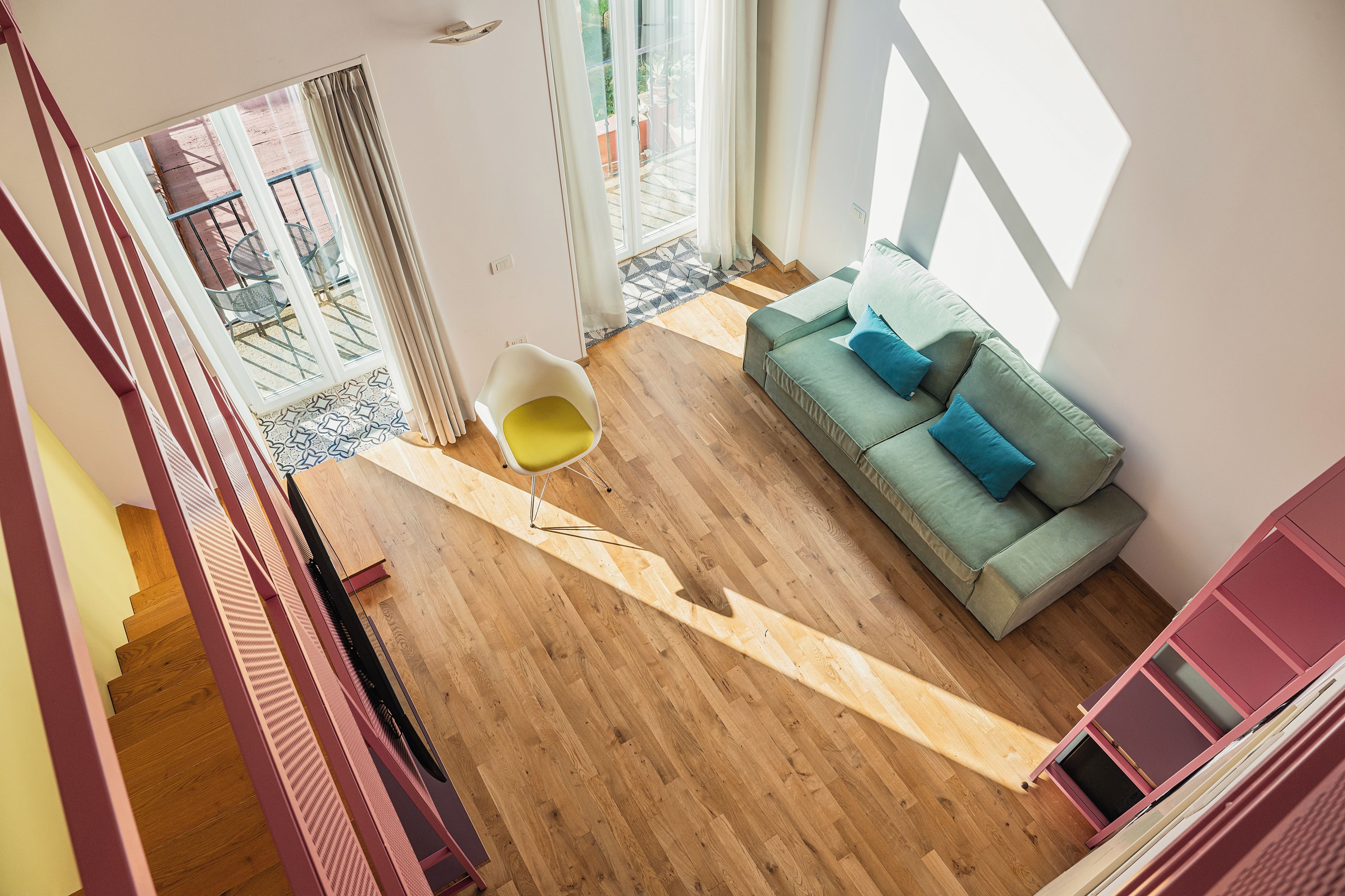 A spacious living area with a wooden floor, a green sofa with blue cushions, a yellow chair, and a glass door leading to an outdoor patio.