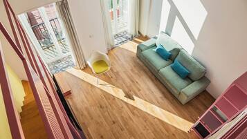 A spacious living area with a wooden floor, a green sofa with blue cushions, a yellow chair, and a glass door leading to an outdoor patio.
