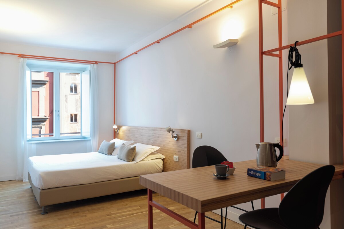 A minimalist hotel room with a bed, a wooden table with a kettle, a chair, and a window with curtains.