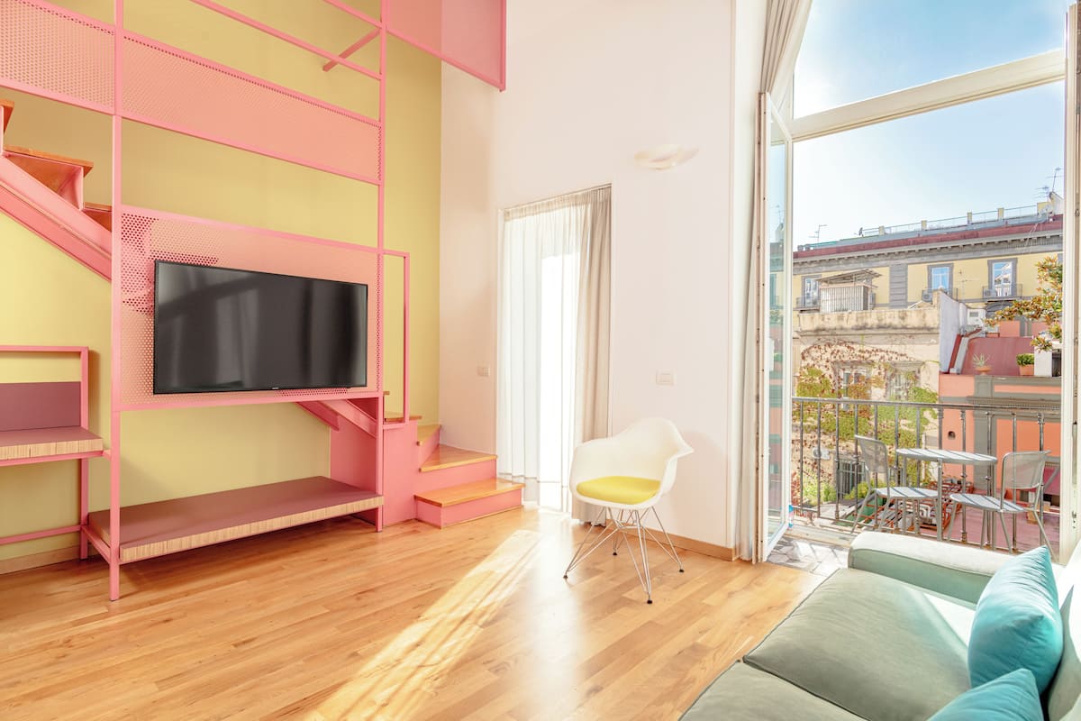 A modern living room with a pink staircase, a flat-screen TV, a white chair, and a balcony with outdoor furniture.
