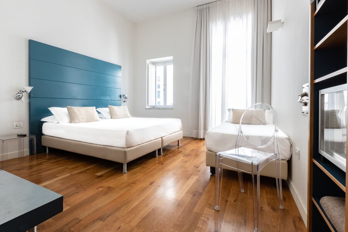 A modern hotel room with a large bed, a small seating area, and a window with curtains.