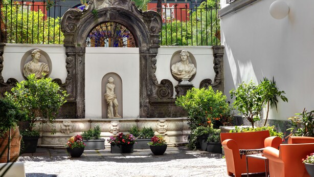 An outdoor courtyard with ornate stone sculptures, potted plants, and orange seating. - Hotel Piazza Bellini & Apartments (Naples)