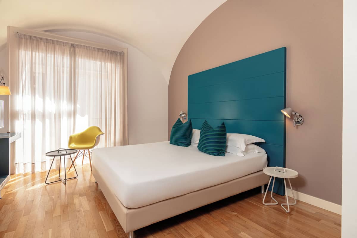 A modern hotel room with a large bed, a teal headboard, a yellow chair, and a small side table.