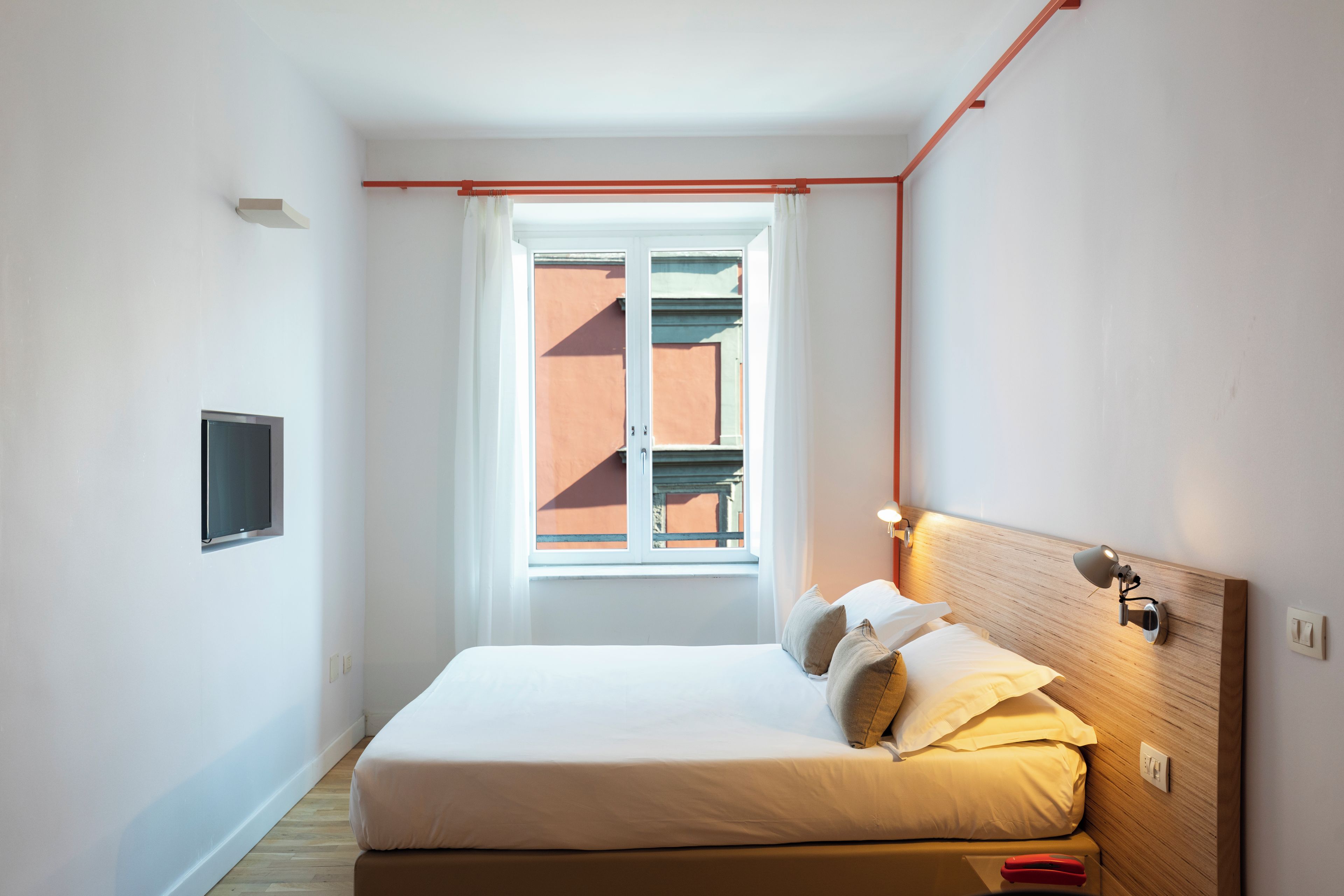 A bedroom with a bed, a window, a wall-mounted TV, and a bedside lamp.