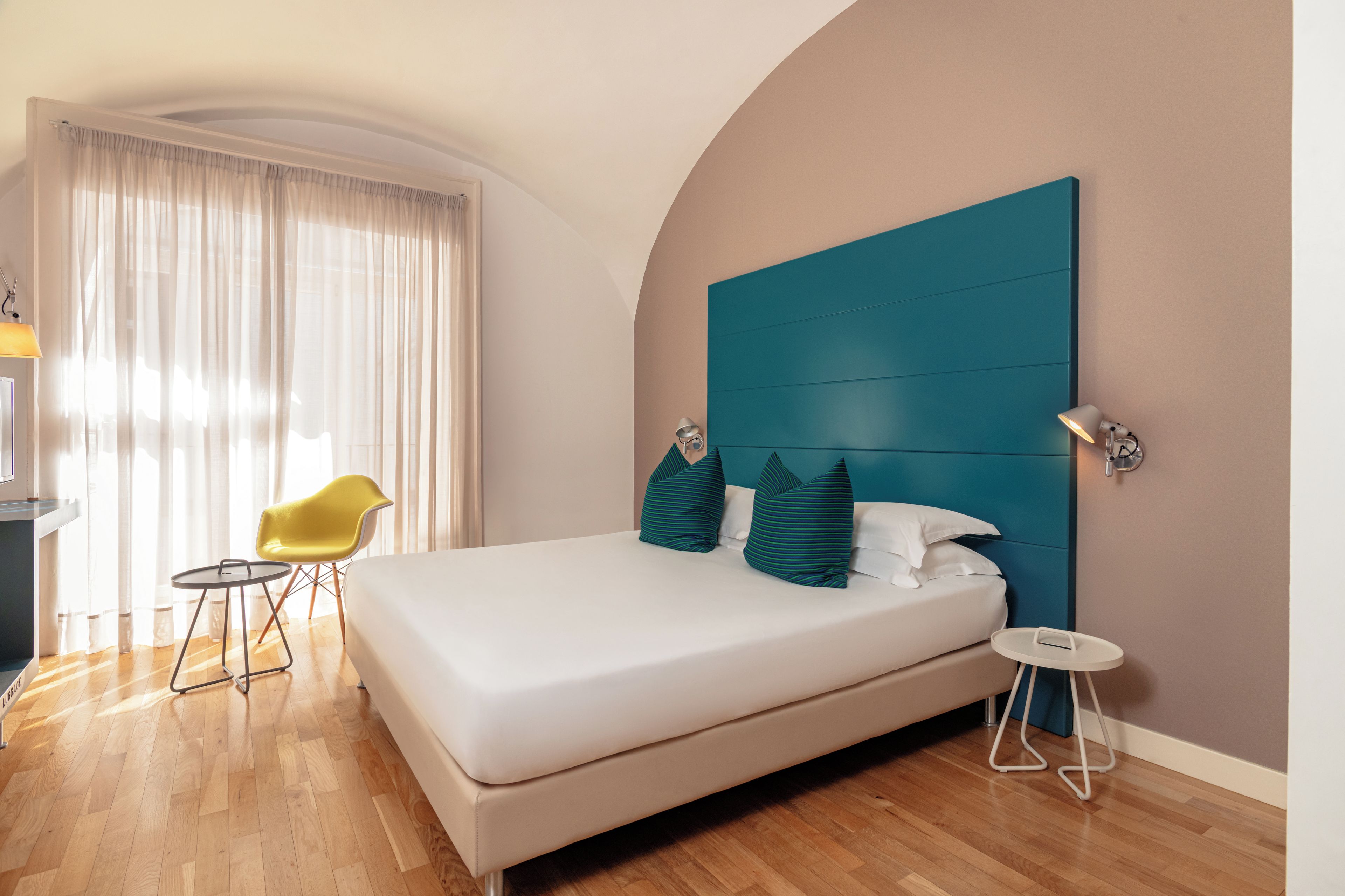 A modern hotel room with a large bed, a teal headboard, a yellow chair, and a small table.