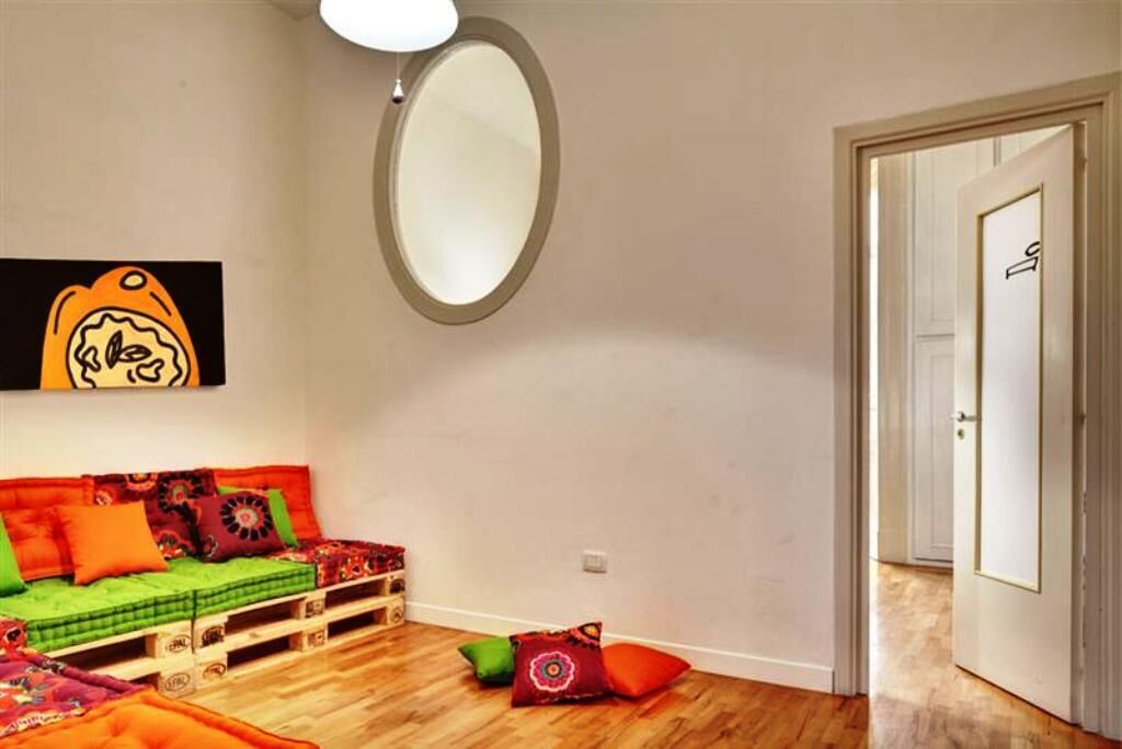 A room with a wooden floor, a green sofa with colorful cushions, a large mirror, and a door with a simple design.