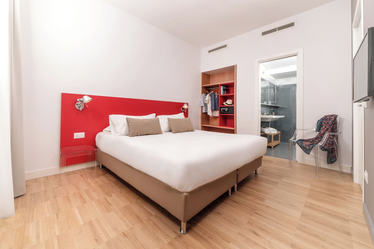 A modern bedroom with a red headboard, a wooden floor, a white bed, a small bedside table, and a wardrobe with a mirror.