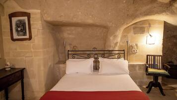 A bedroom with a stone wall, a bed with white linens, a red blanket, a wooden nightstand, a chair, and a picture frame on the wall.