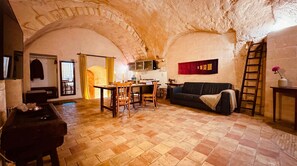 Un intérieur rustique avec un plafond voûté en pierre, un coin repas avec des meubles en bois, un canapé noir et une échelle adossée au mur.