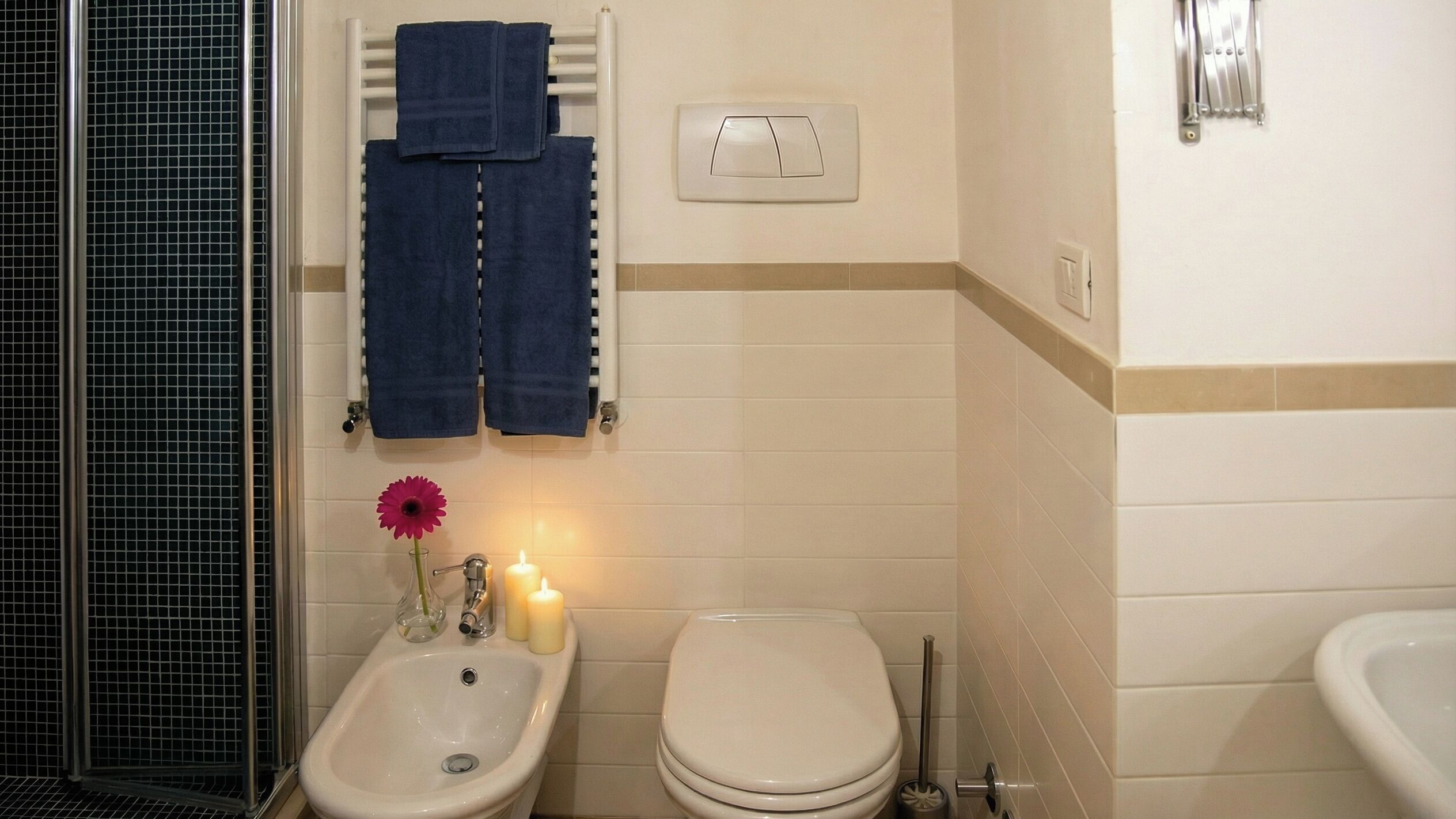 A bathroom with a bidet, toilet, and sink. There are towels hanging on a rack, a flower vase, and two lit candles.