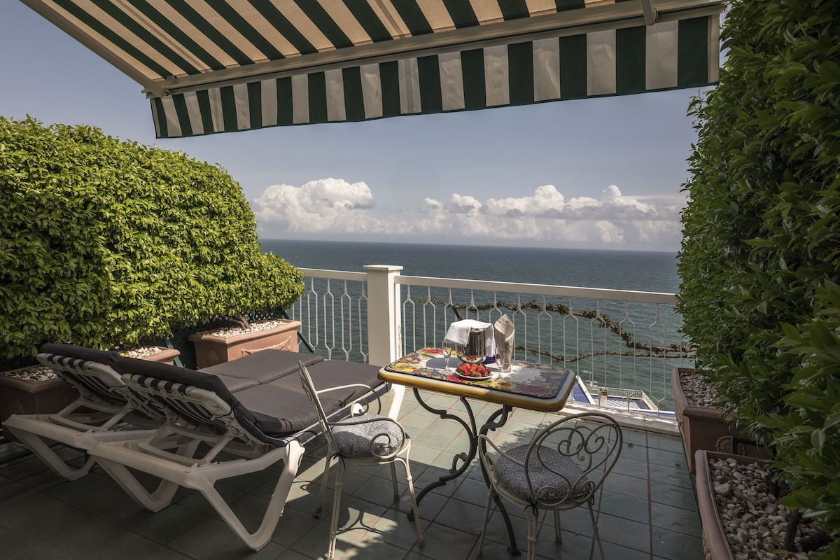 Un balcon avec vue sur la mer, une table recouverte d’une nappe et deux fauteuils de jardin.
