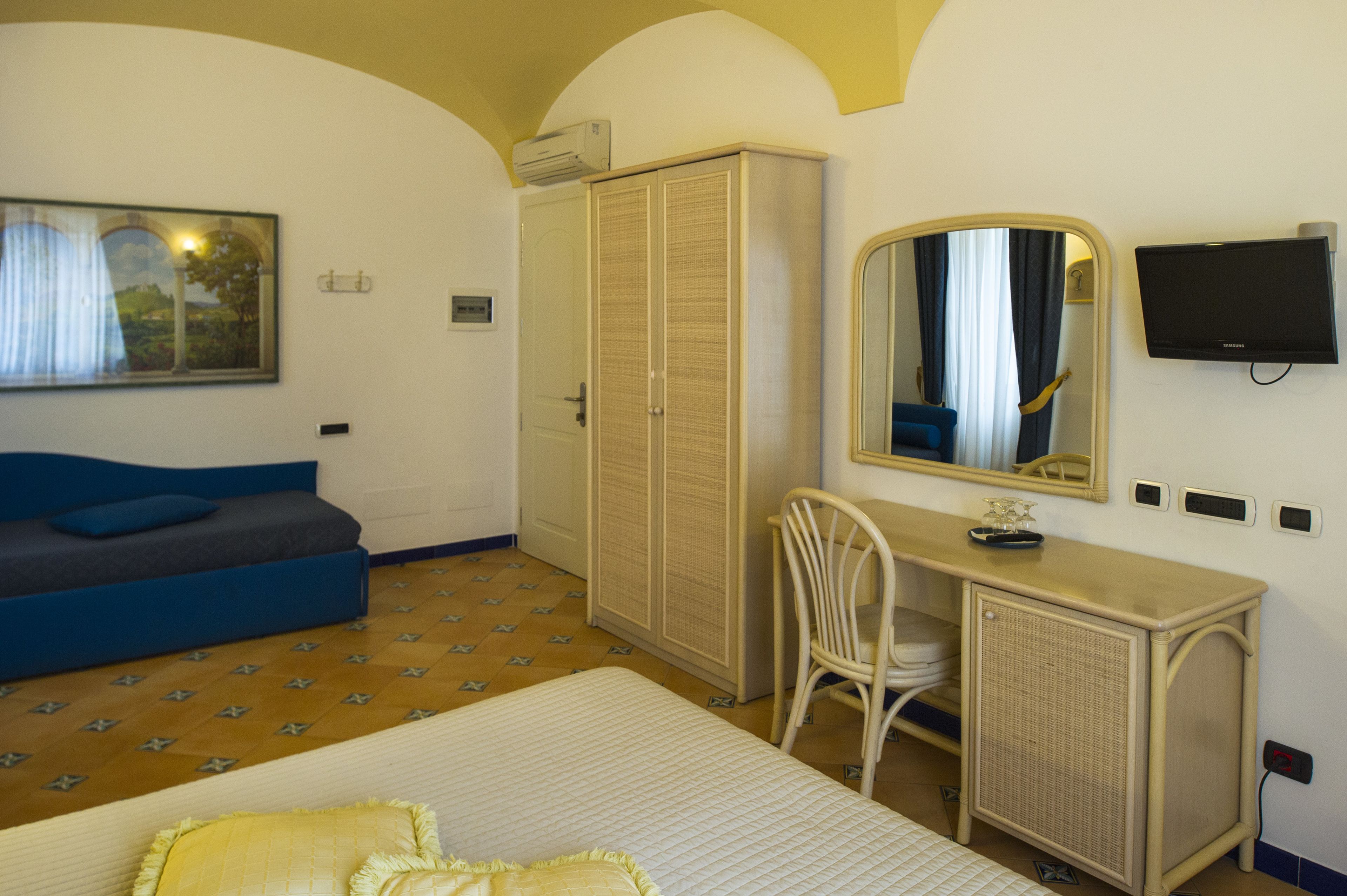 A hotel room with a bed, a blue sofa, a desk with a mirror, a wardrobe, and a television.