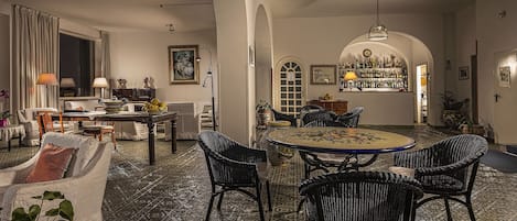 A spacious living room with a dining area, featuring a patterned carpet, wicker furniture, and a bar area with a wine collection.
