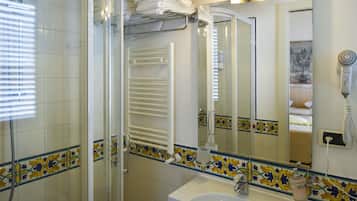 Un baño con cabina de ducha de vidrio, un lavamanos blanco, un perchero con toallas dobladas, un espejo con borde de azulejos decorativos y un secador de pelo en la pared.