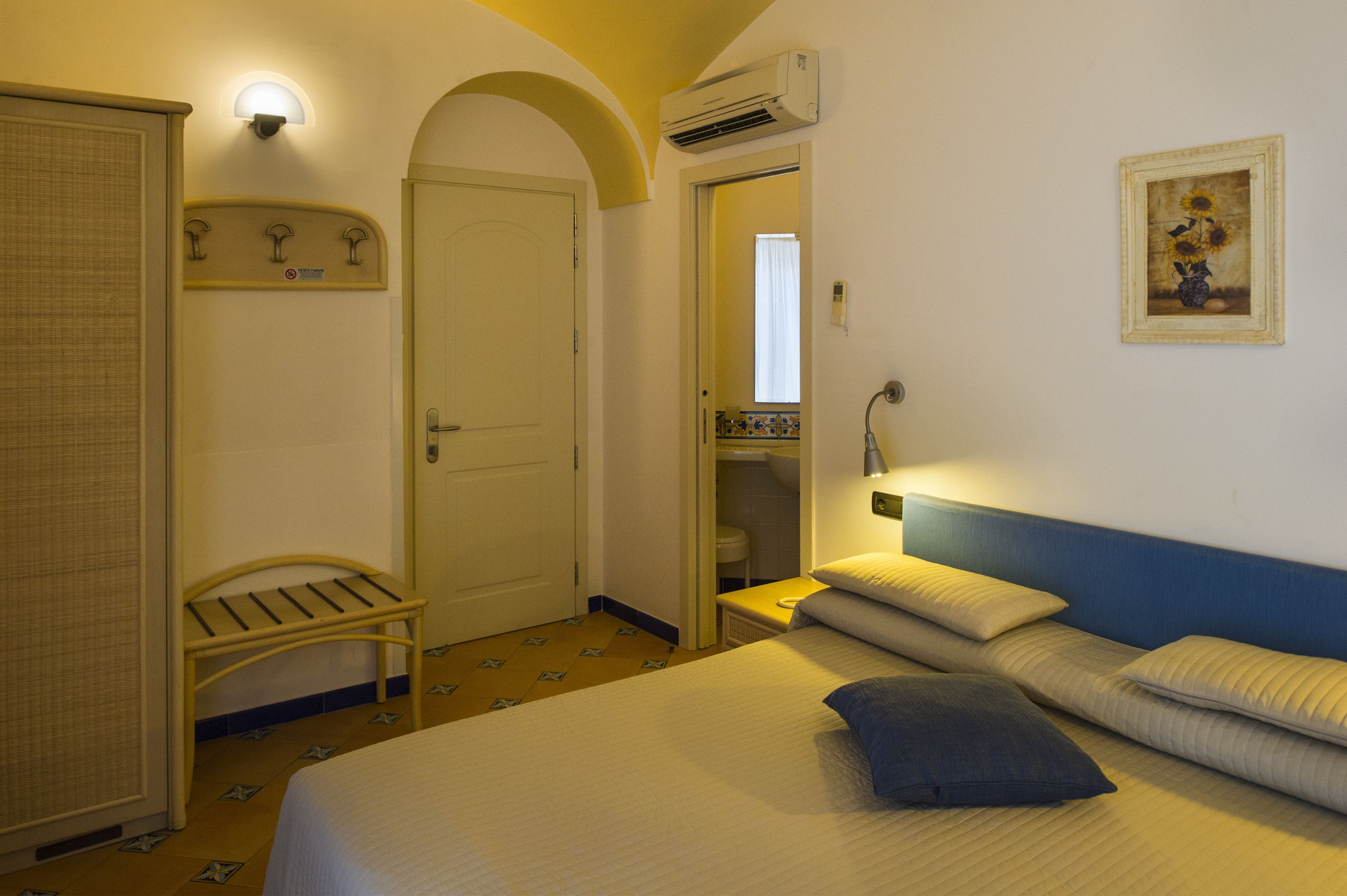 A hotel room with a bed, a bench, a door, and a wall-mounted air conditioner.