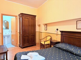 Habitación de hotel con armario de madera, mesita de noche, silla, cama con edredón estampado, lámpara de noche y un cuadro en la pared.