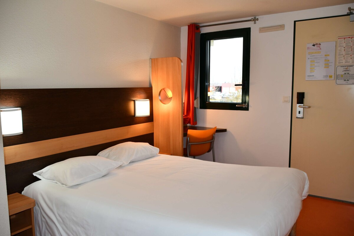 A hotel room with a bed, a desk, a chair, a window with curtains, and a door with a sign.