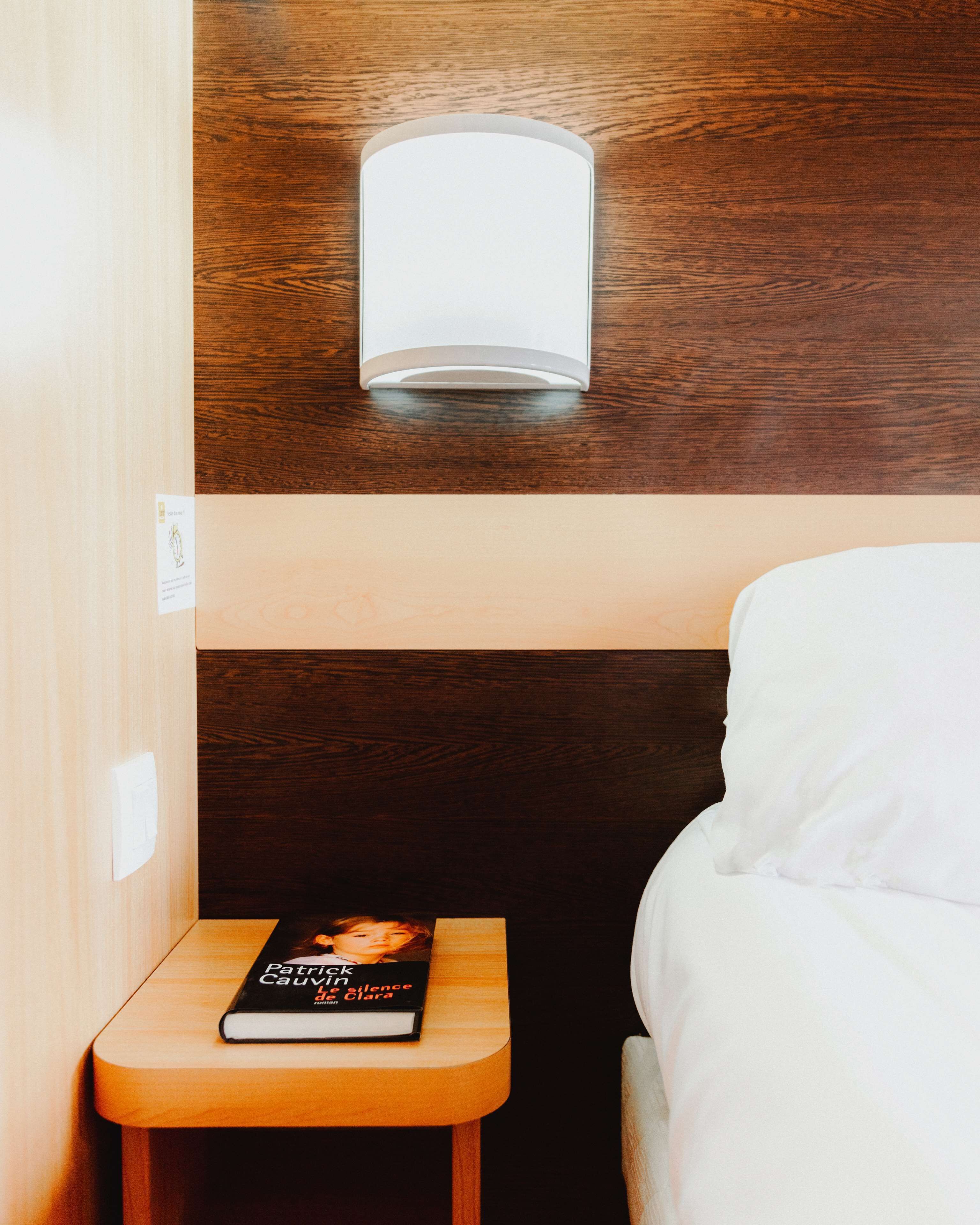 A neatly made bed with white linens, a bedside table with a book titled "Patricia & Silence Clara," and a wall-mounted light fixture.
