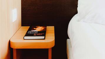 A neatly made bed with white linens, a bedside table with a book titled "Patricia & Silence Clara," and a wall-mounted light fixture.