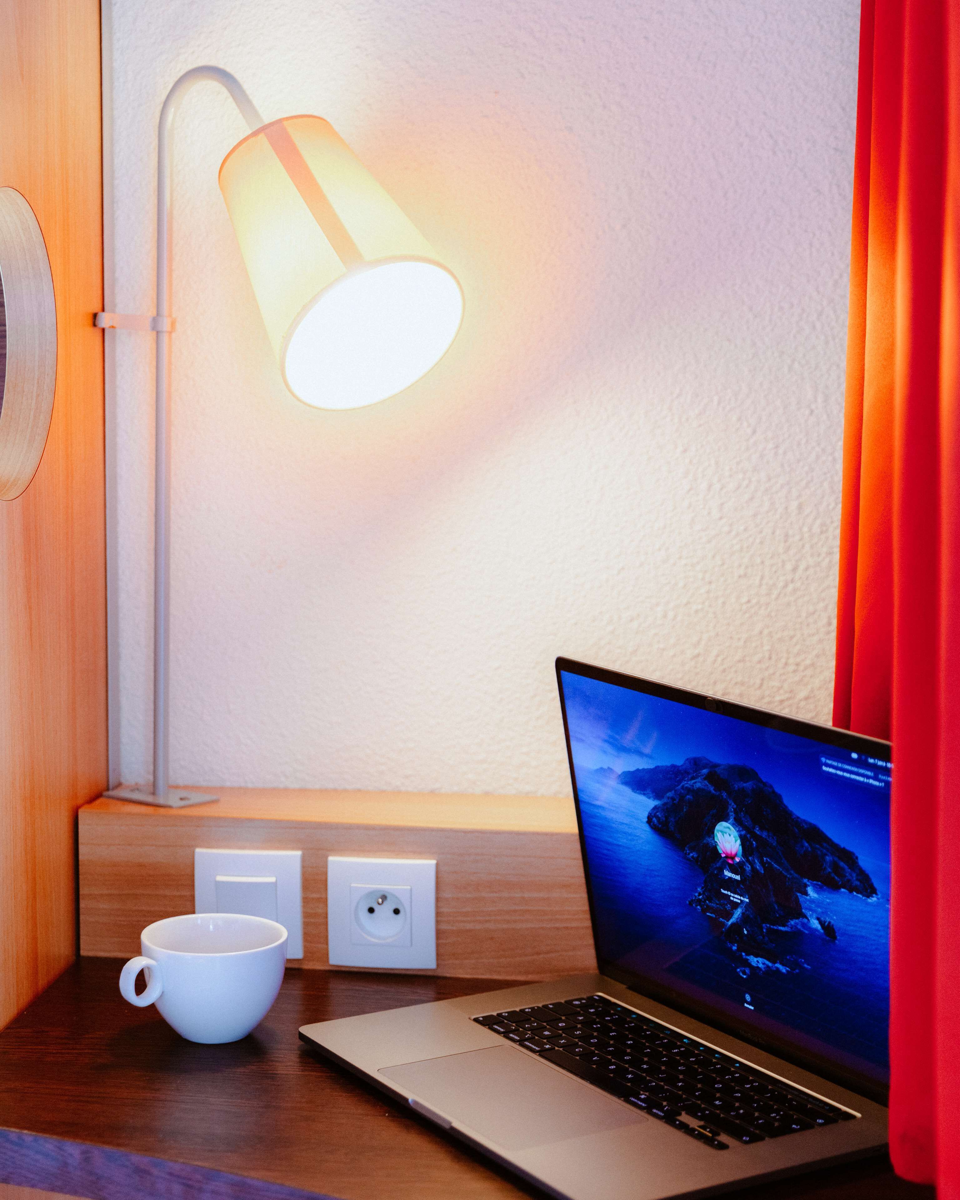 Un ordinateur portable posé sur un bureau en bois, avec une tasse blanche, une lampe fixée au mur et une prise de courant.