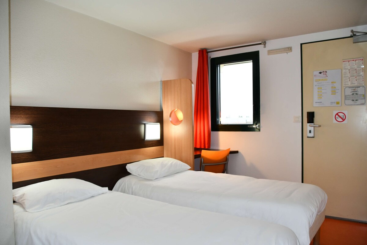 A hotel room with two beds, a wooden headboard, a window with red curtains, and a door with a no-smoking sign.