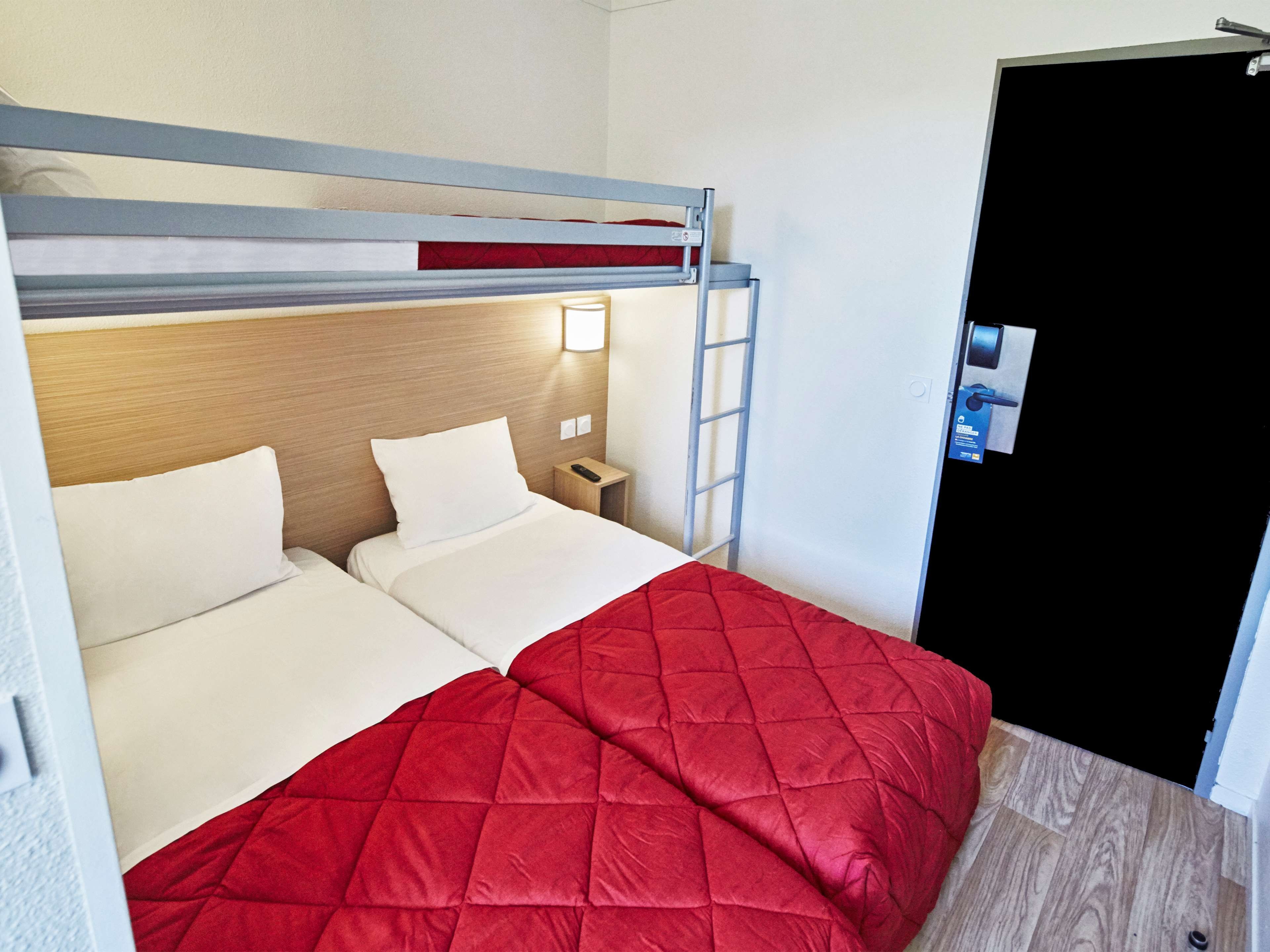 Una cama litera con ropa de cama blanca y una colcha acolchada roja. Hay una mesita de noche con un control remoto y una lámpara fijada a la pared. Una escalera está apoyada contra la pared.