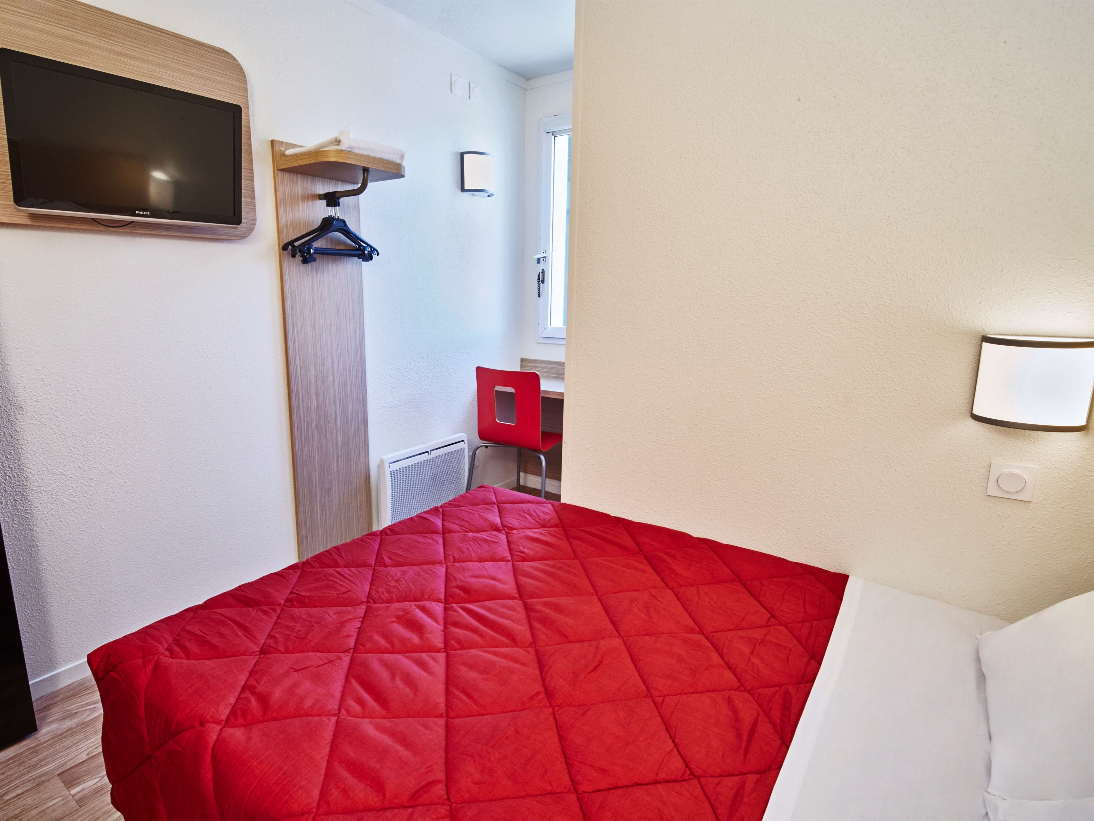 A hotel room with a bed, a television mounted on the wall, a red chair, a small table, and a red cabinet.