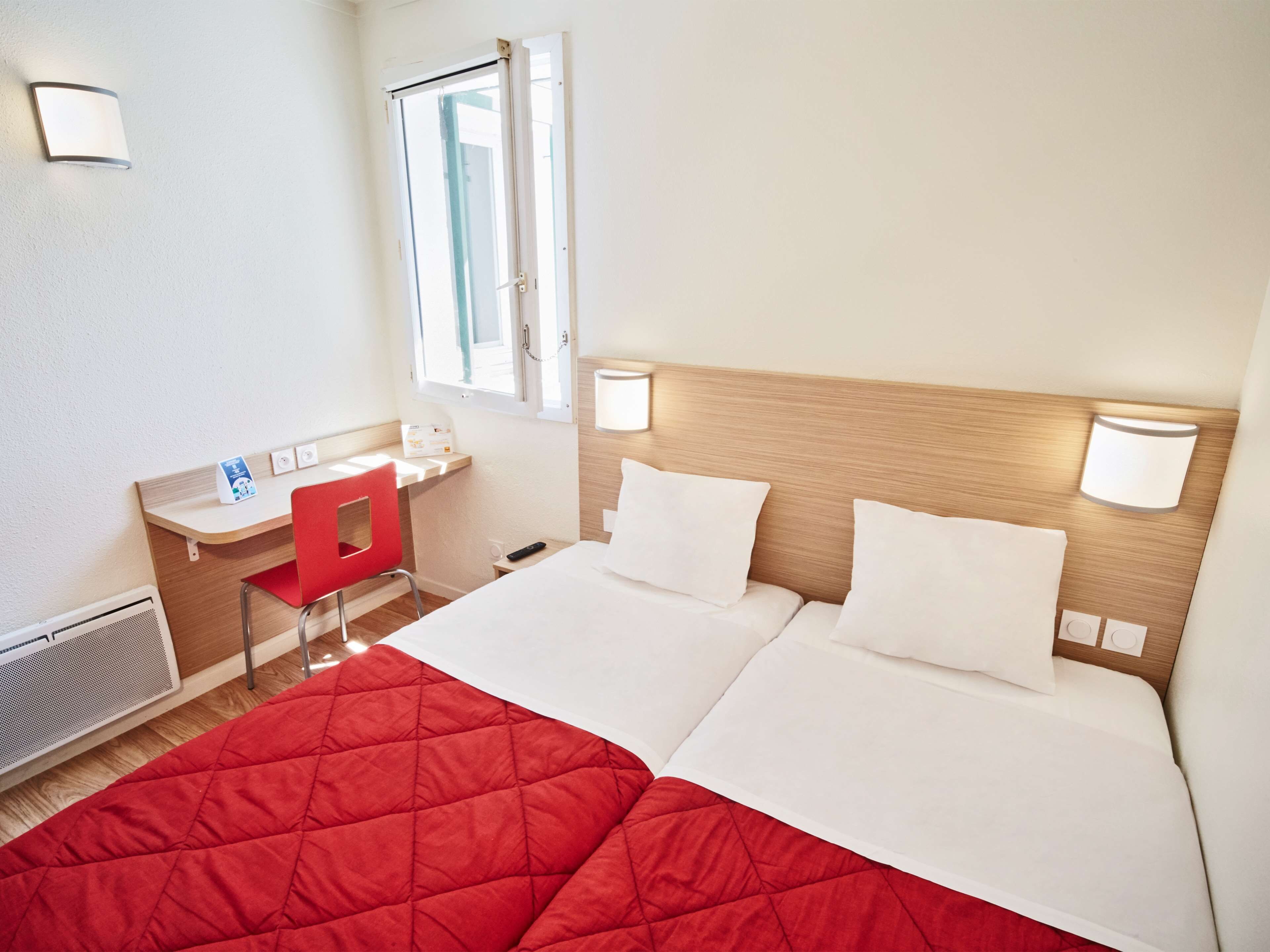 A hotel room with a bed, a desk, a chair, a window, and a wall-mounted lamp.