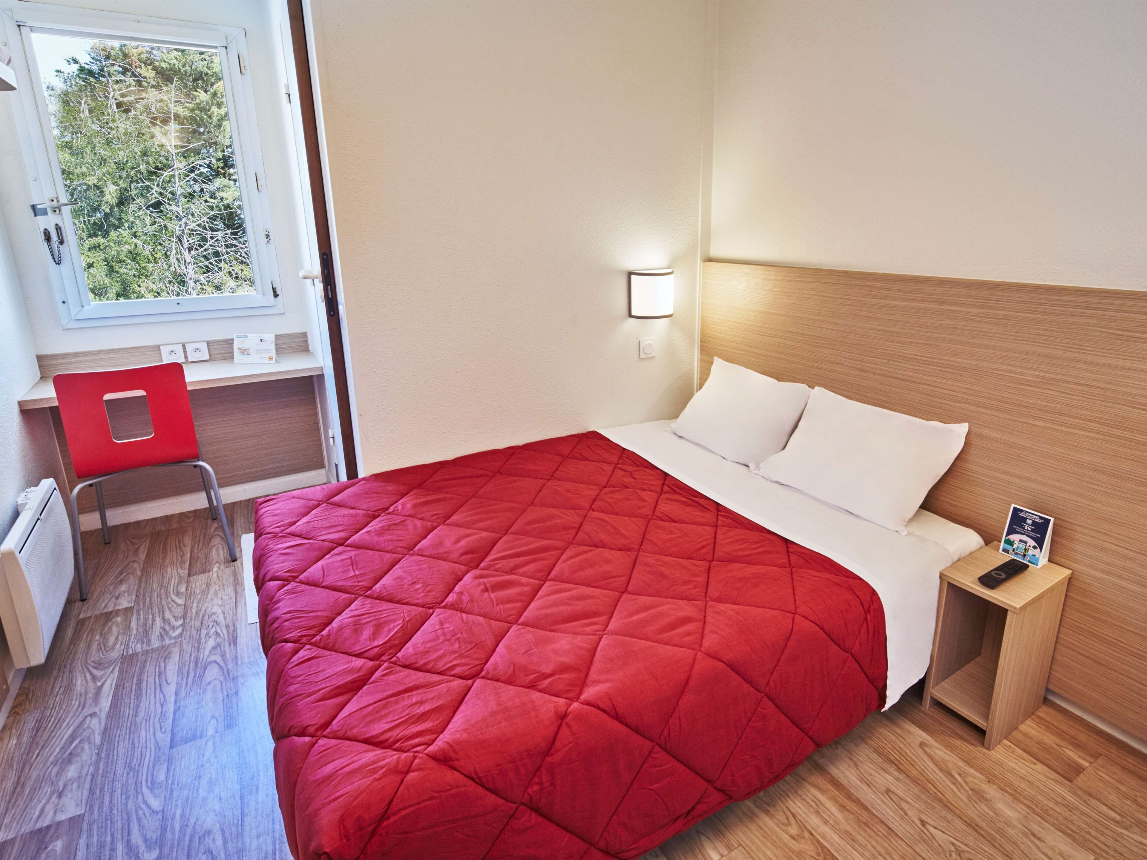 A hotel room with a bed, a desk, a chair, a nightstand, and a window with a view of trees.
