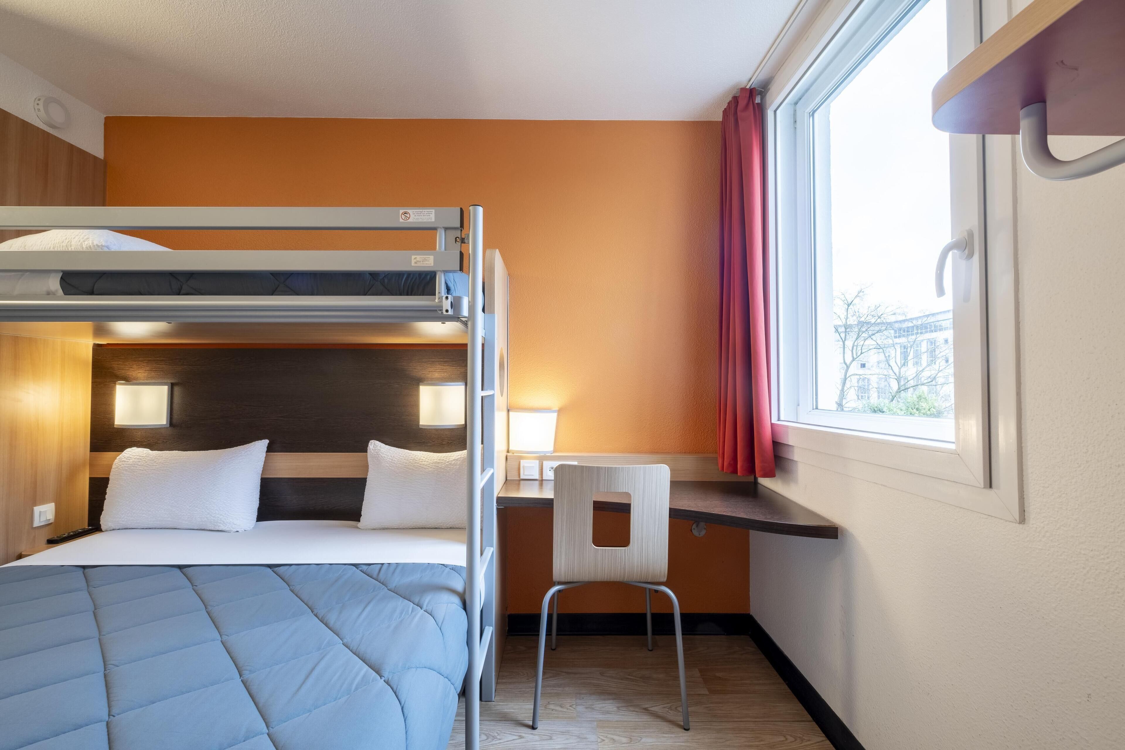 A hotel room with a bunk bed, a desk with a chair, a window with curtains, and a lamp.