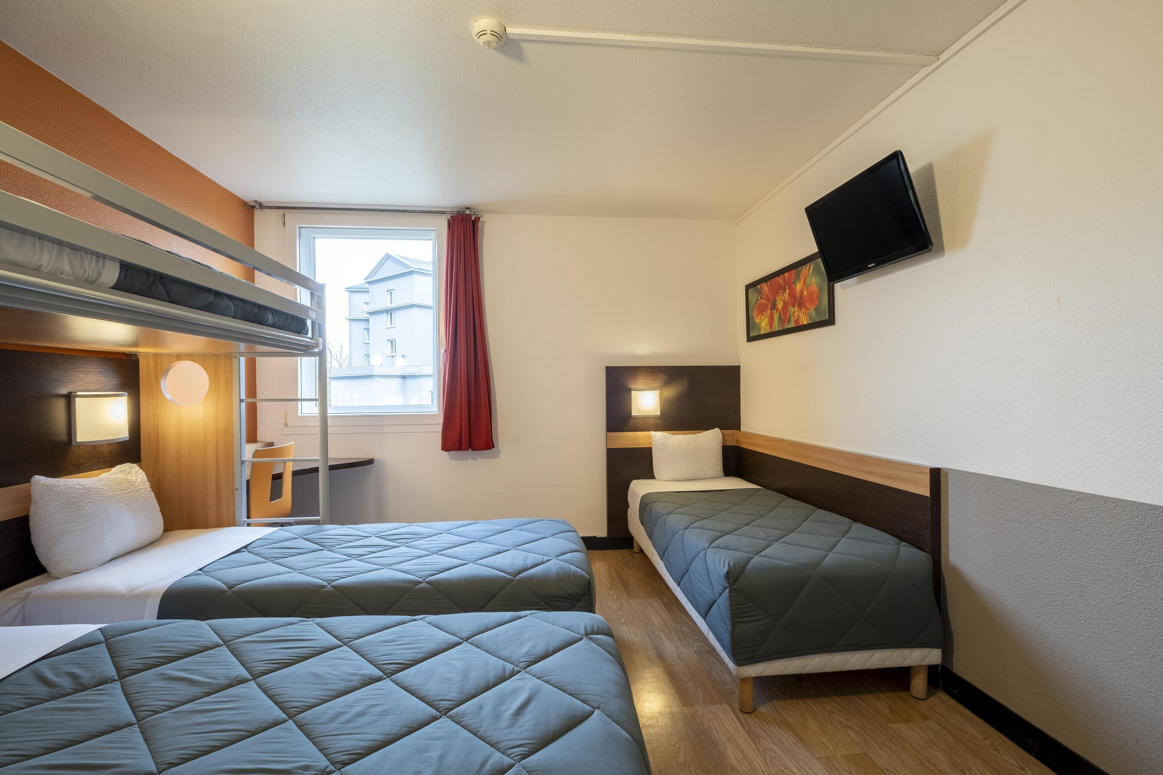 A hotel room with two beds, a desk, a chair, a TV, and a window with a red curtain.