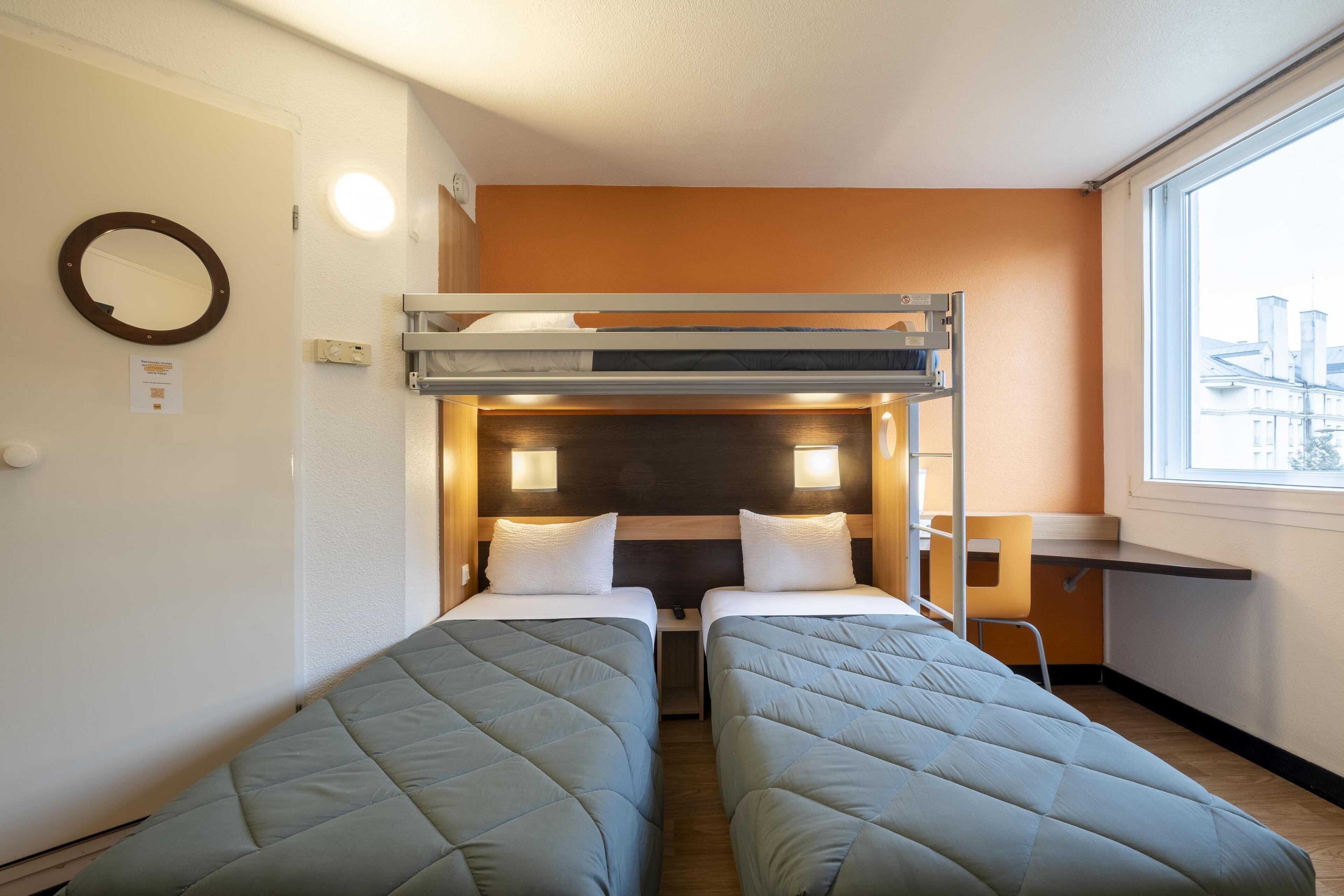 A compact hotel room with two single beds, a bunk bed, a small desk, and a window with a city view.