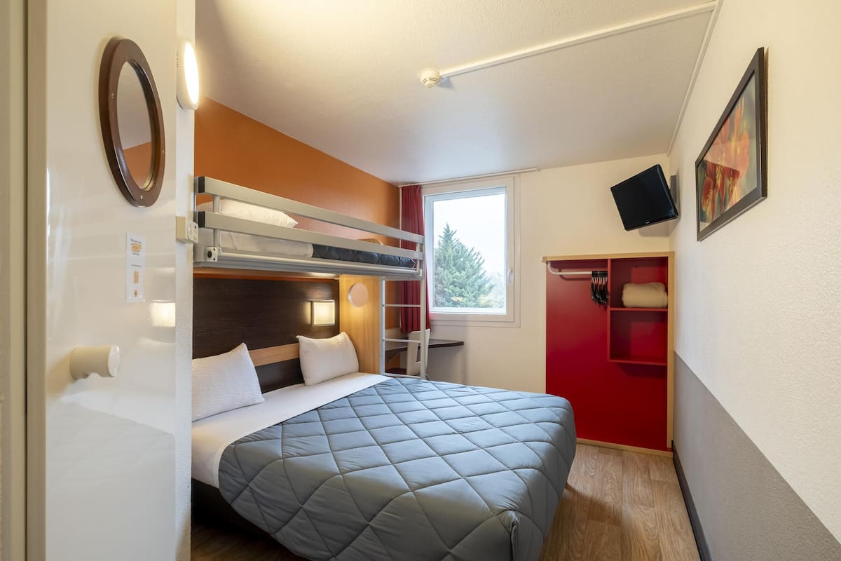 Ein kleines Hotelzimmer mit Etagenbett, Schreibtisch, Stuhl, Fernseher und Fenster mit Blick auf Bäume.