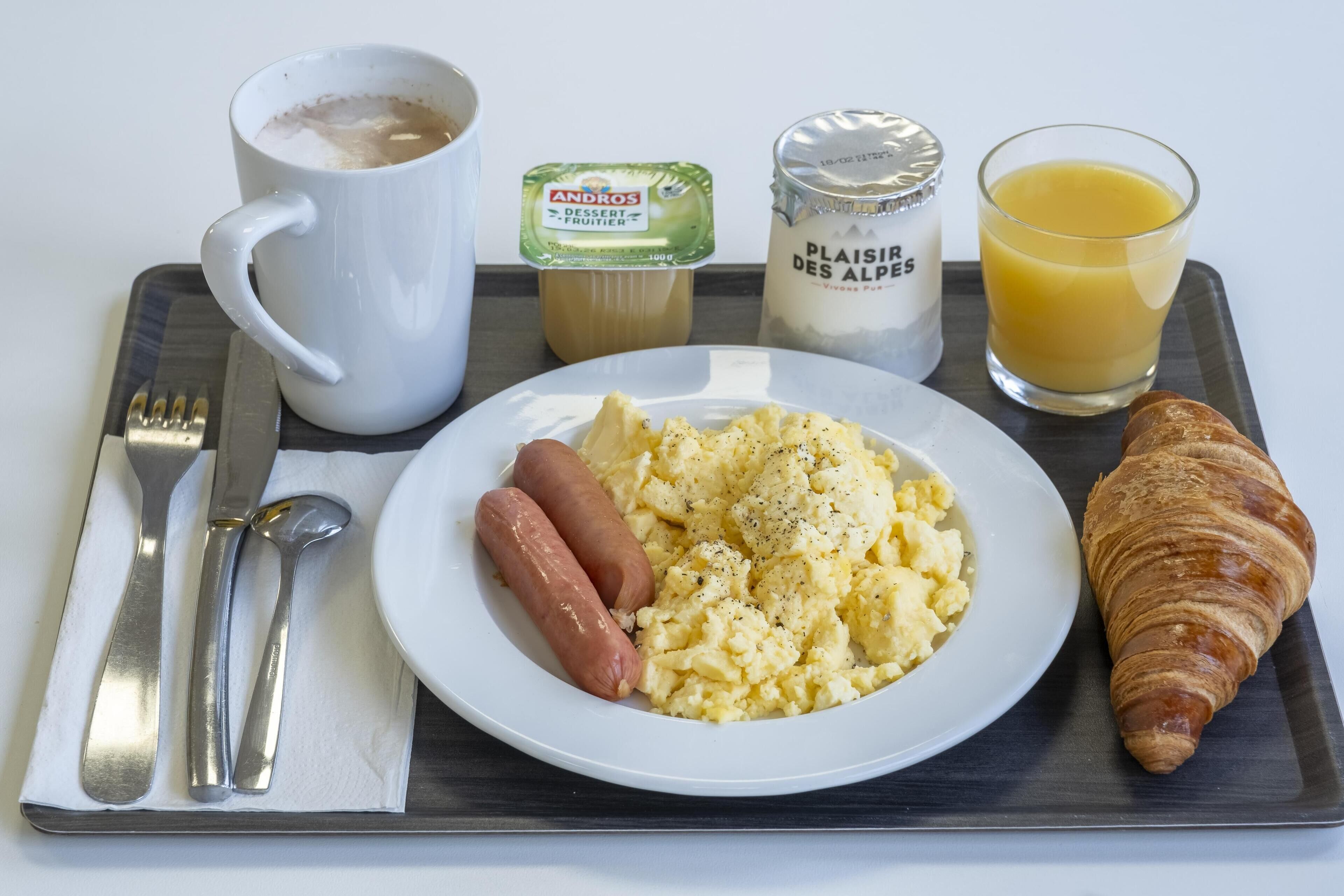 A breakfast tray with scrambled eggs, sausages, a croissant, a glass of orange juice, a coffee mug, a container of yogurt, and condiments.