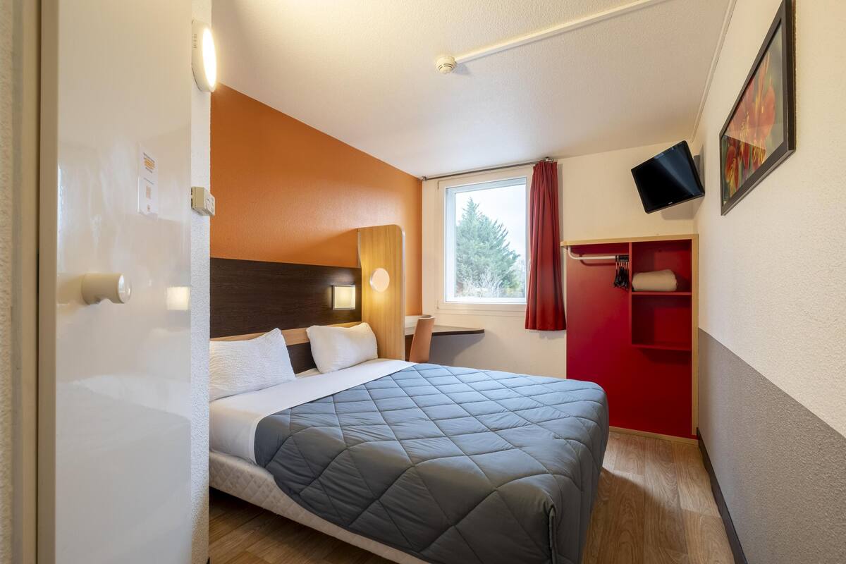 Ein kleines Hotelzimmer mit Bett, Schreibtisch, einem Fenster mit Vorhängen, einem Wand-Fernseher und einem roten Kleiderschrank.