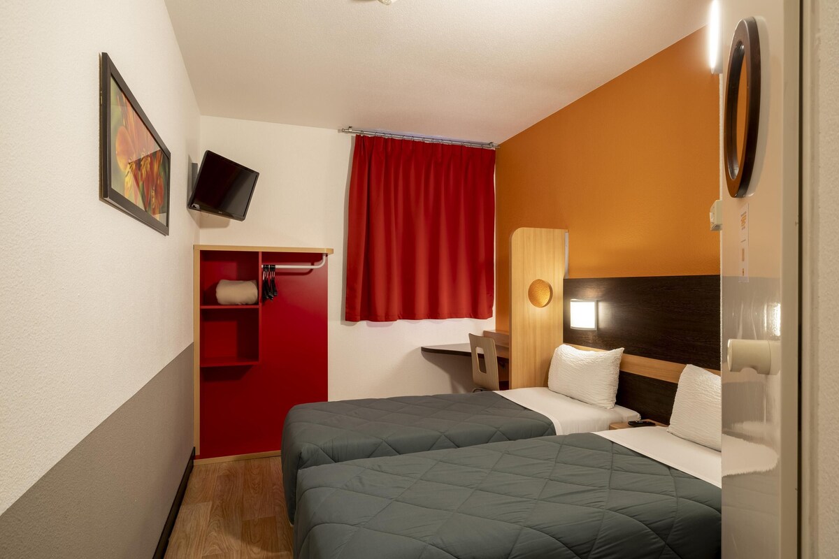 A hotel room with two beds, a desk, a chair, a TV, and a red curtain.