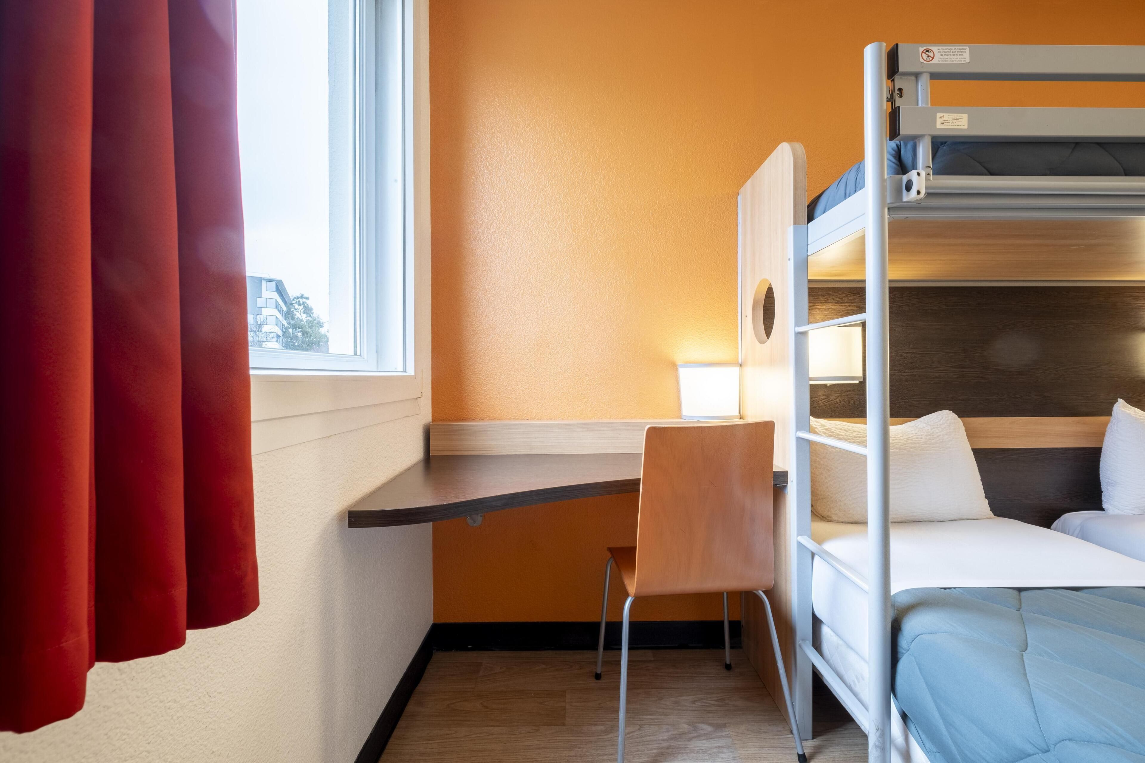 A bunk bed room with a desk and chair, a window with curtains, and a view of buildings outside.