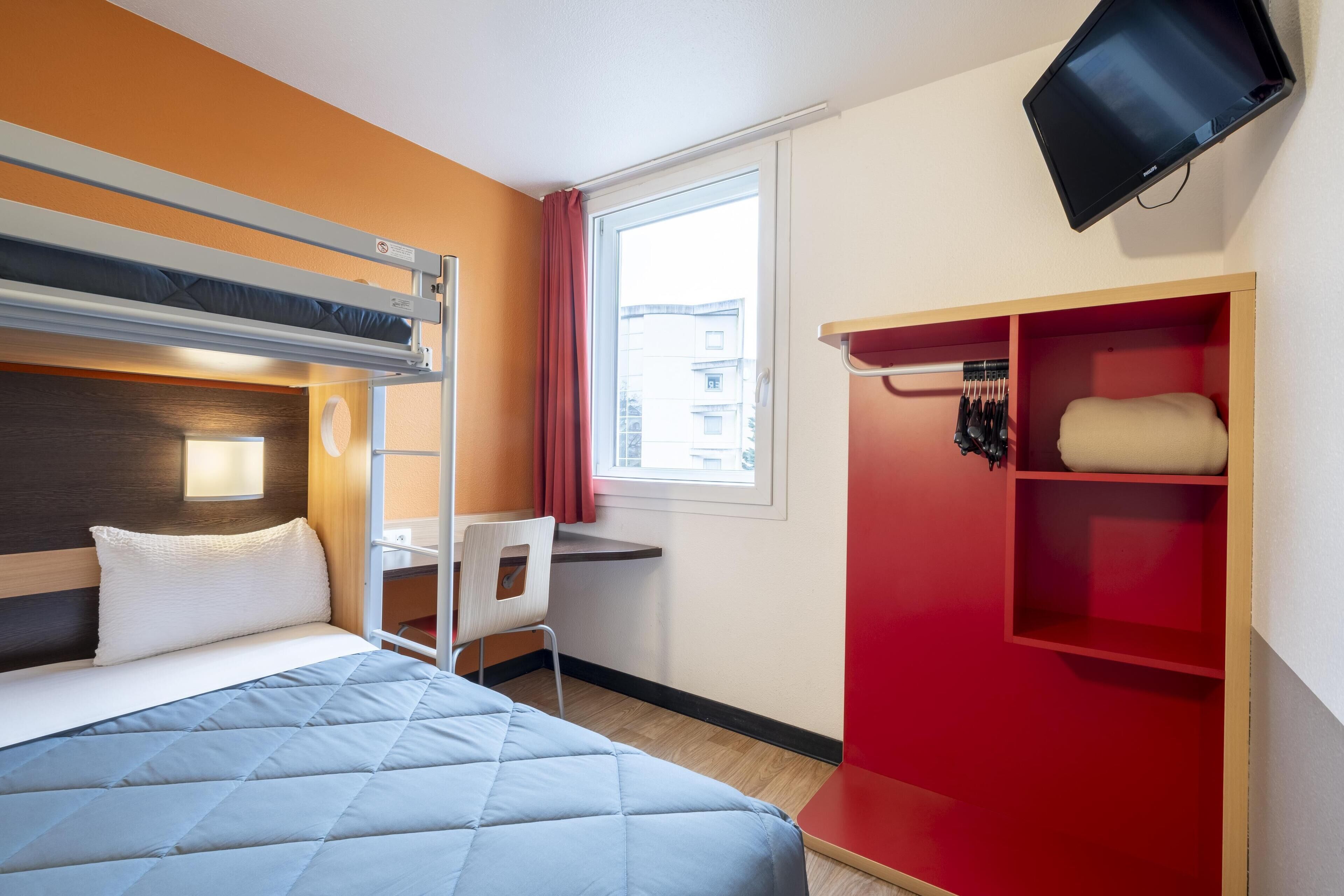 A hotel room with a bunk bed, a desk, a chair, a TV, and a red storage unit.