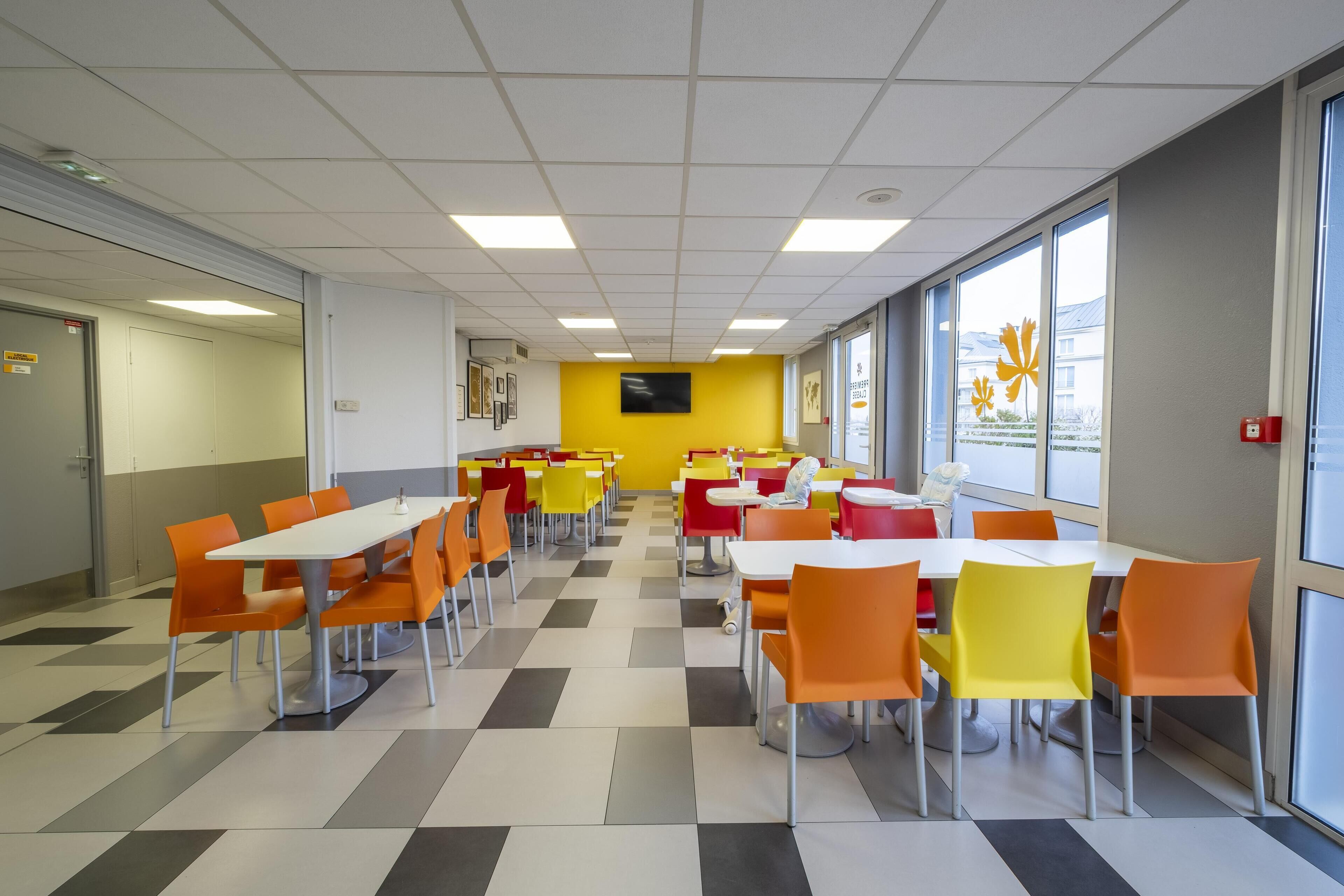 Una cafetería con varias mesas y sillas, una pared de acento amarilla y grandes ventanales con vista a edificios.