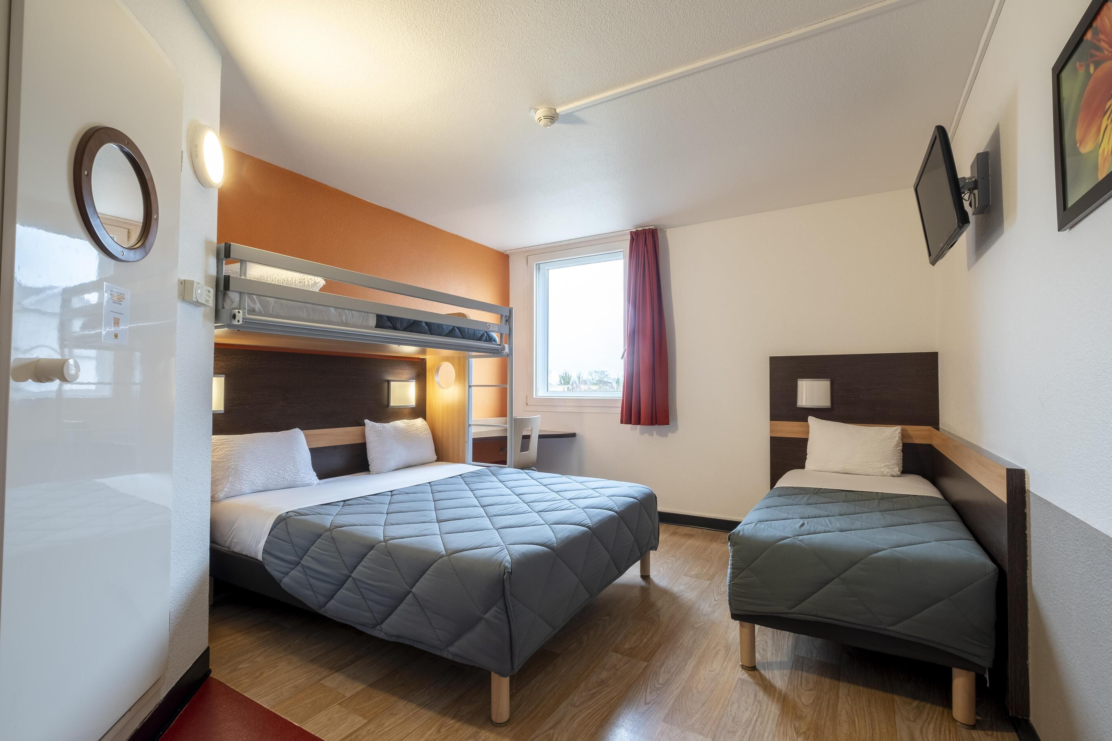 A room with two beds, a bunk bed, a window with curtains, a round mirror, and a flat-screen TV mounted on the wall.