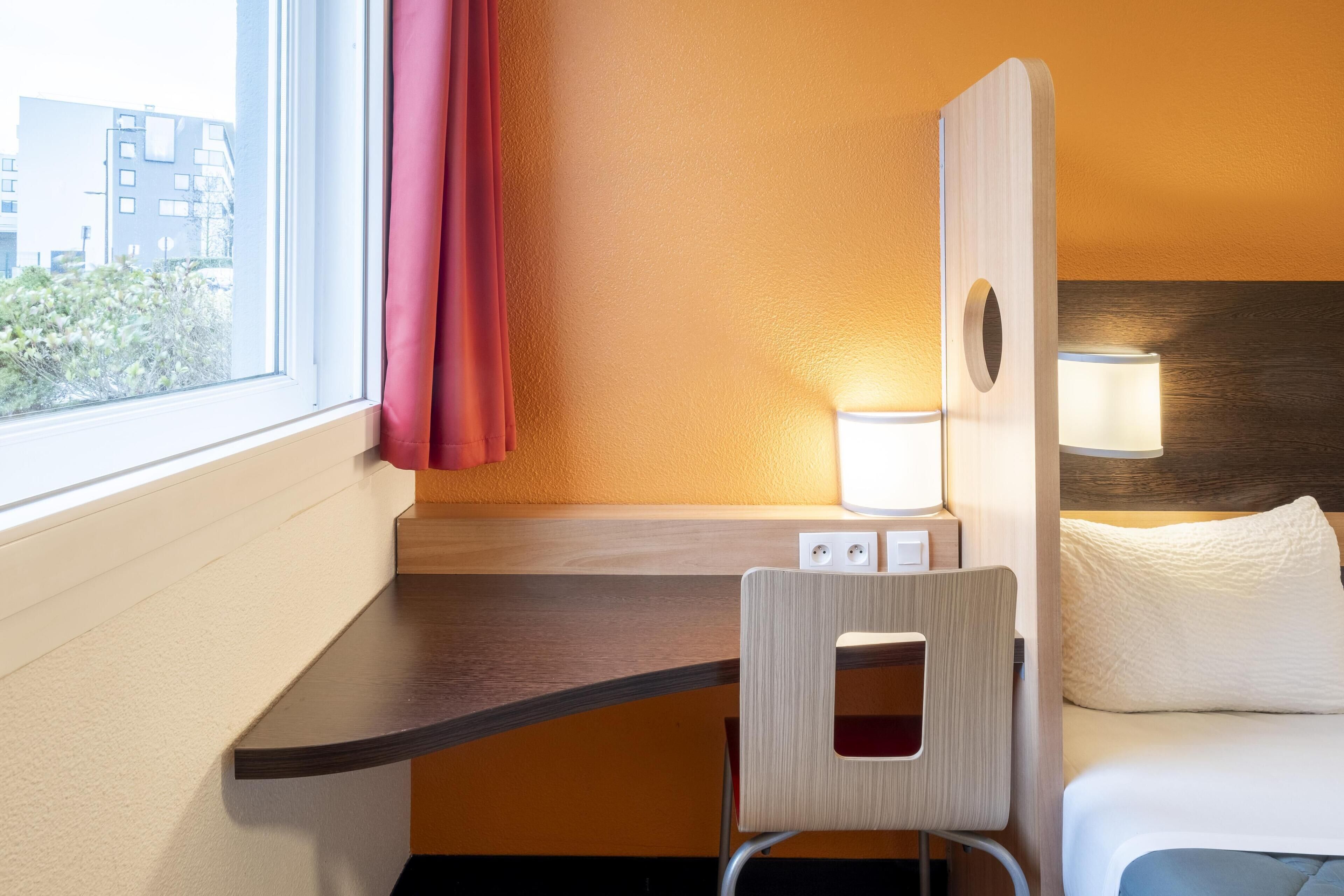 A hotel room with a bed, a desk, a chair, a window with a curtain, and a lamp.