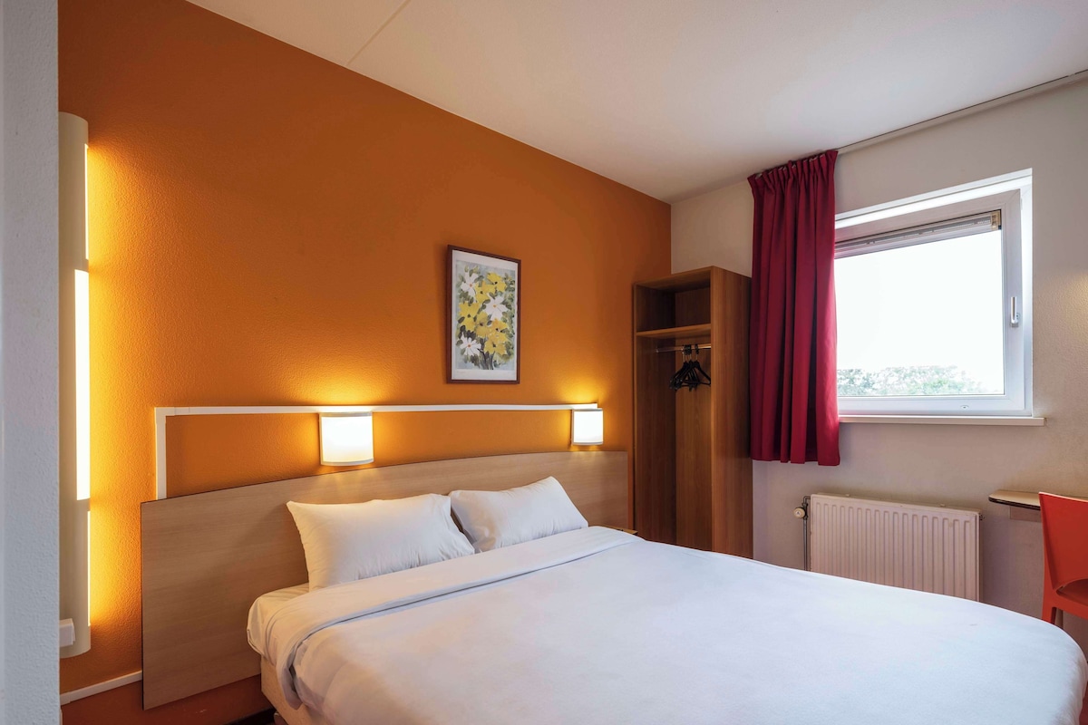 Une chambre d’hôtel avec un lit, des lampes de chevet, une fenêtre avec des rideaux, un radiateur et une petite table.