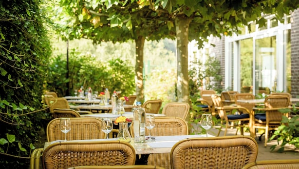 Een terras met rieten tafels en stoelen, omgeven door planten en een bakstenen muur.