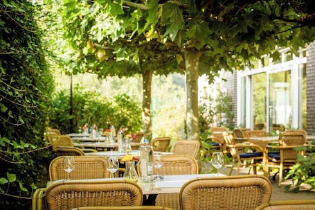An outdoor dining area with wicker tables and chairs, surrounded by greenery and a brick wall.