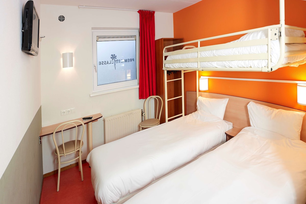 Une chambre d’hôtel avec deux lits, un bureau, une chaise, un radiateur et une fenêtre avec des rideaux.