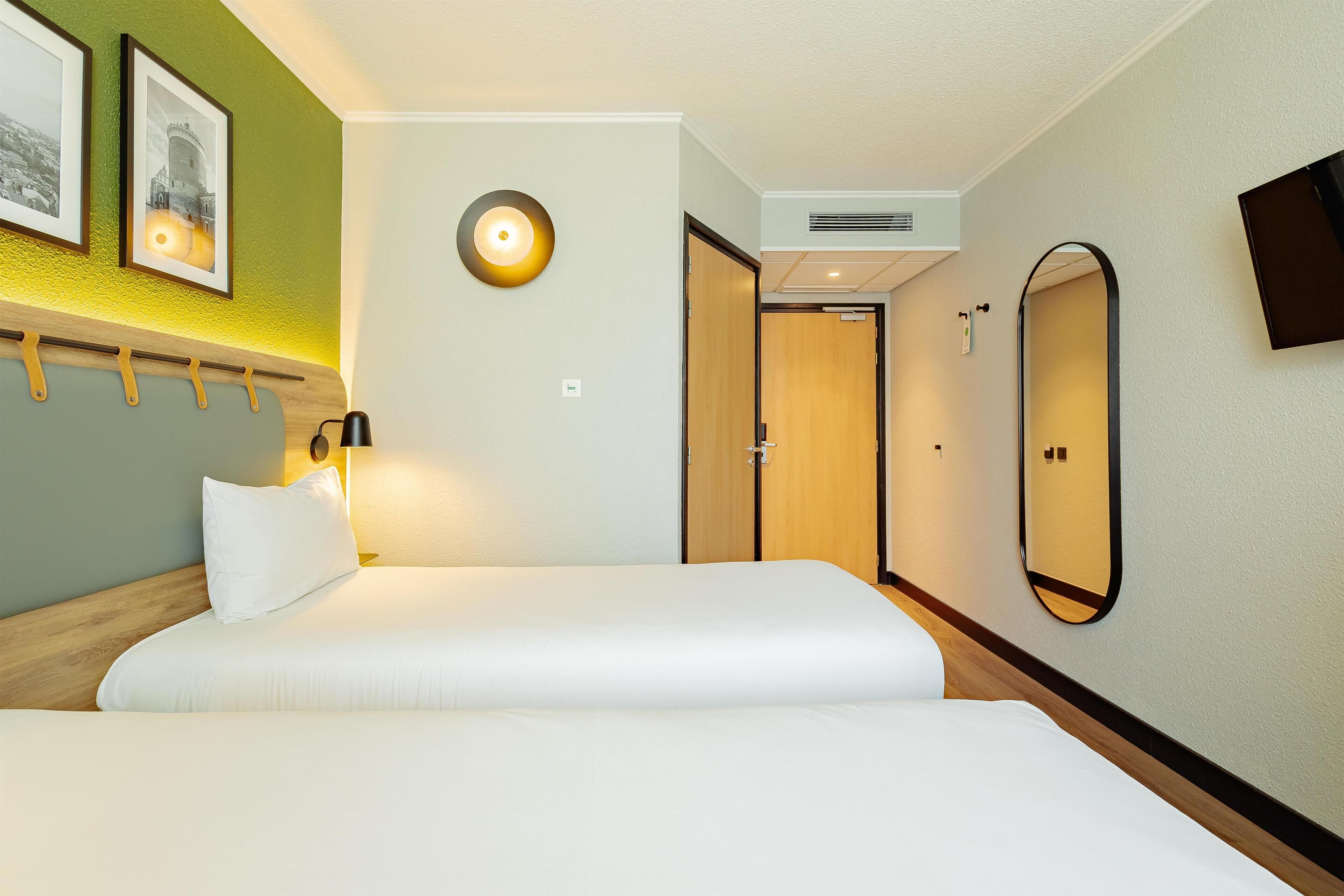 A hotel room with a bed, a wall-mounted TV, a mirror, and a door leading to another room.