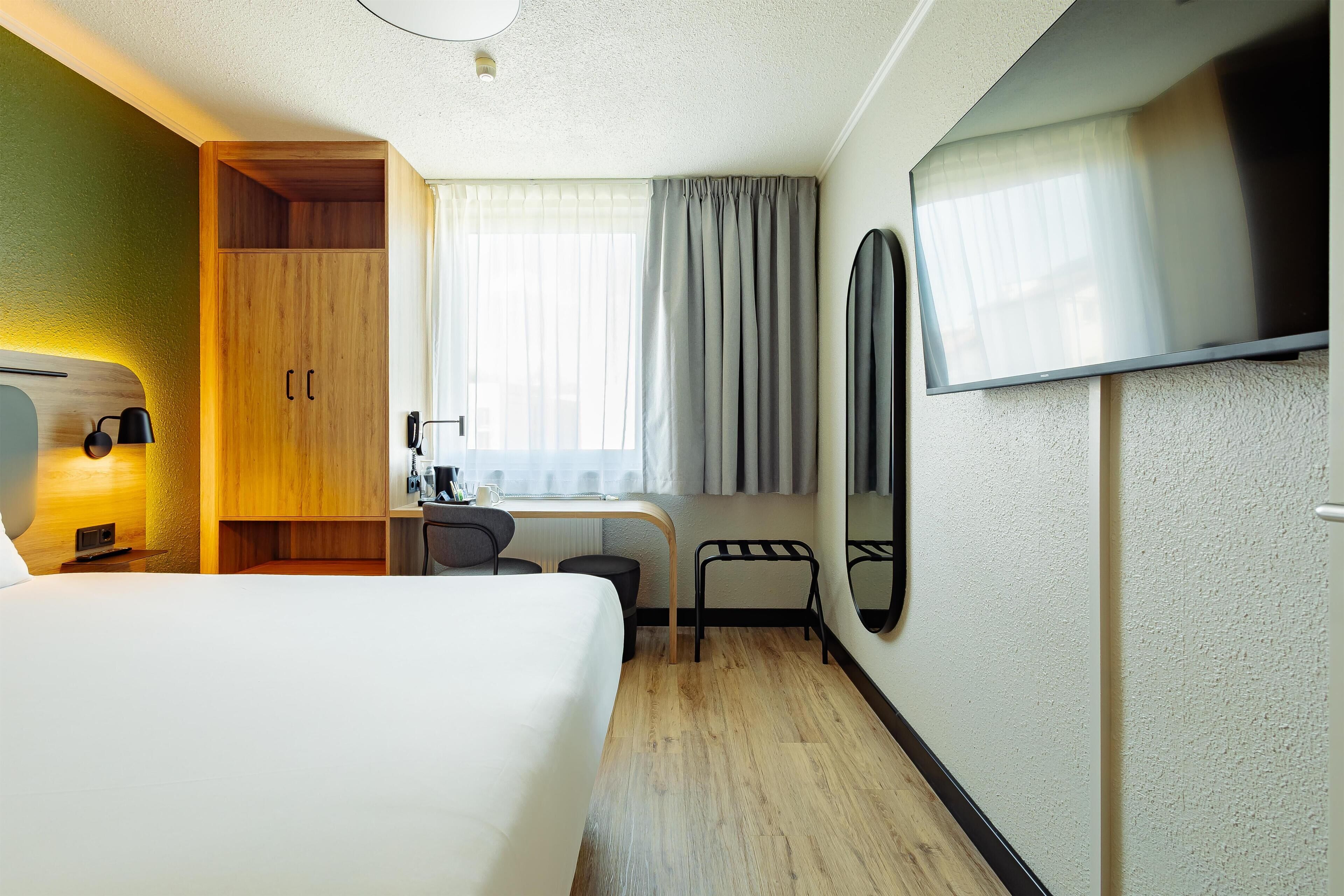 A hotel room with a bed, desk, chair, wardrobe, and a TV.